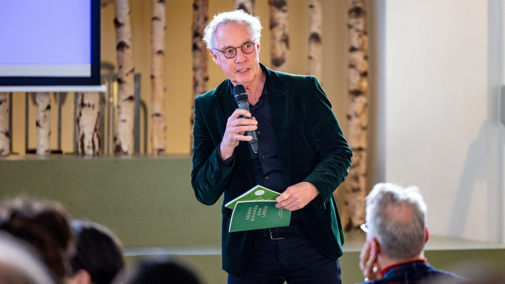 Robert Koch met microfoon en boekjes in de hand voor een publiek